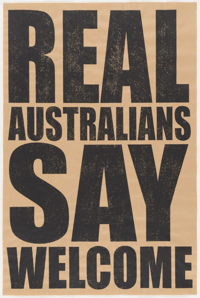 A brown screen printed poster with black capital letters that read 'REAL AUSTRALIANS SAY WELCOME'.